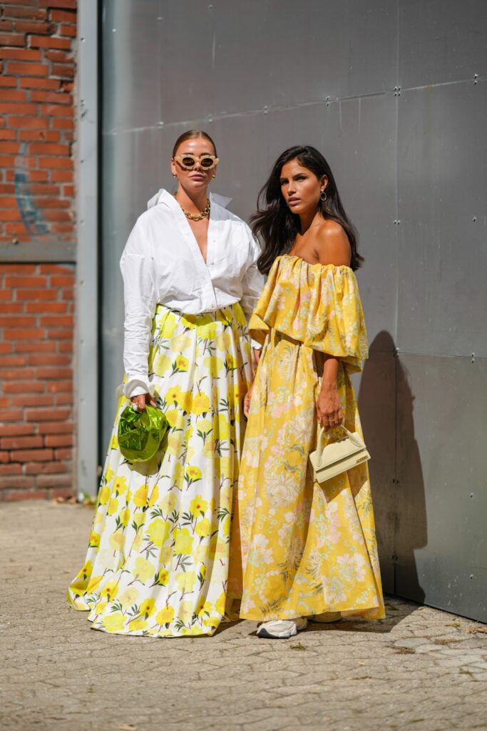 Две жени облечени в пролетни стилизации - едната с бяла широка риза и жълта дълга пола на цветя и друата с жълта флорална рокля и маратонки